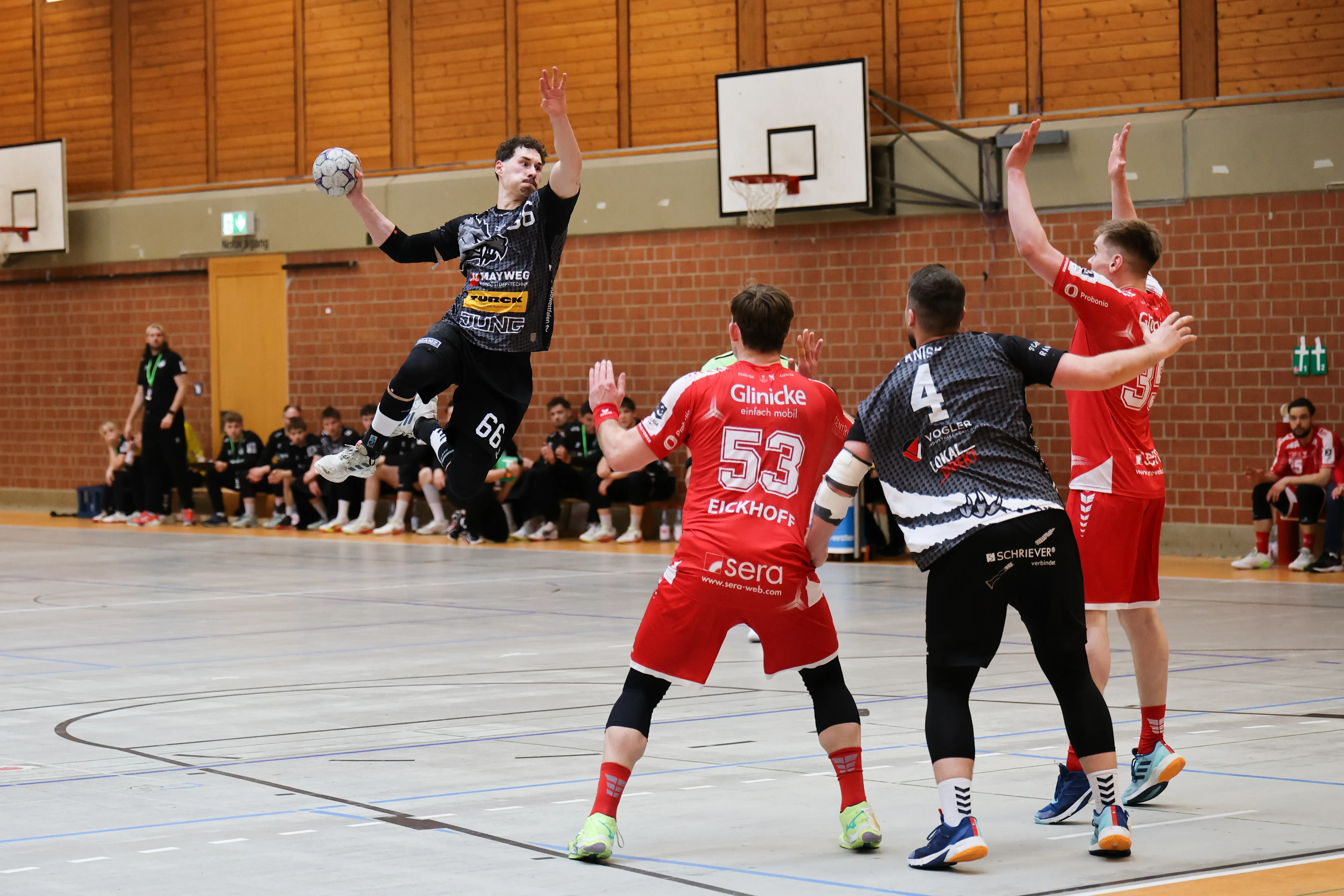 Handballspieler springt in einer Sporthalle hoch, um den Ball zu werfen, während Gegenspieler versuchen, ihn zu blocken. Im Hintergrund stehen weitere Spieler und Zuschauer.