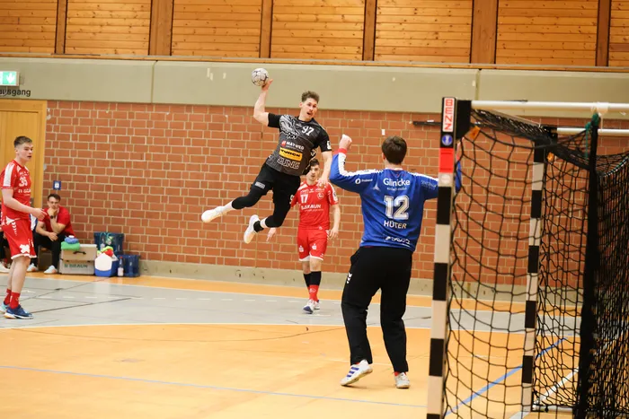 Handballspieler in dunklen und roten Trikots im Spiel. Ein Spieler springt zum Wurf, während ein Torwart versucht, den Ball zu blocken.