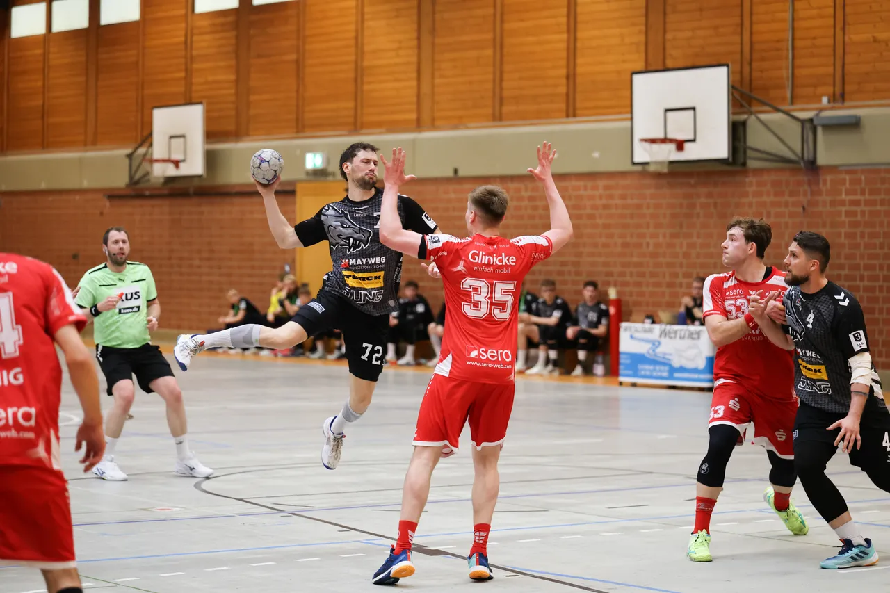 Handballspieler springen in einer Sporthalle um den Ball. Ein Spieler im schwarzen Trikot mit der Nummer 72 wirft, während ein Spieler in Rot mit der Nummer 35 blockt.