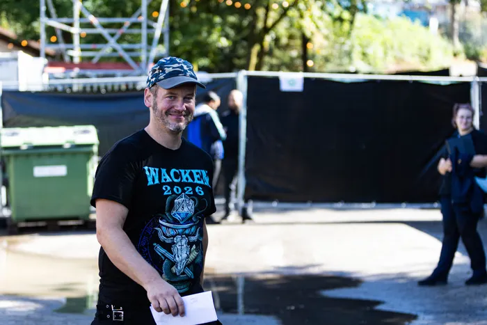 Ein lächelnder Mann mit Camouflage-Kappe und einem schwarzen T-Shirt mit der Aufschrift „WACKEN 2026“ hält ein weißes Papier.