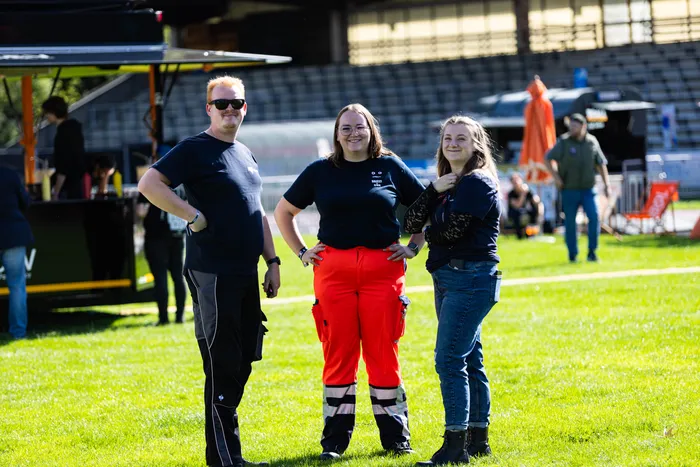 Drei lächelnde Personen stehen auf einer sonnigen Wiese vor Festivalständen; eine Frau trägt eine auffällige rote Arbeitshose.