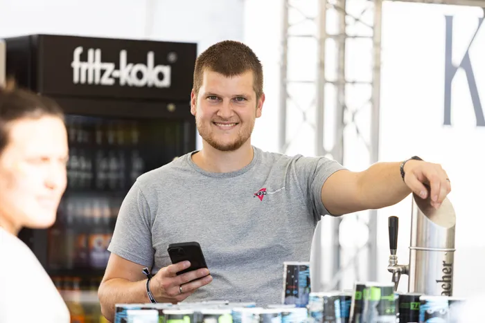 Ein lächelnder Mann in einem grauen T-Shirt bedient an einem Stand einen Zapfhahn und hält ein Smartphone. Im Hintergrund steht ein Fritz-Kola-Kühlschrank.