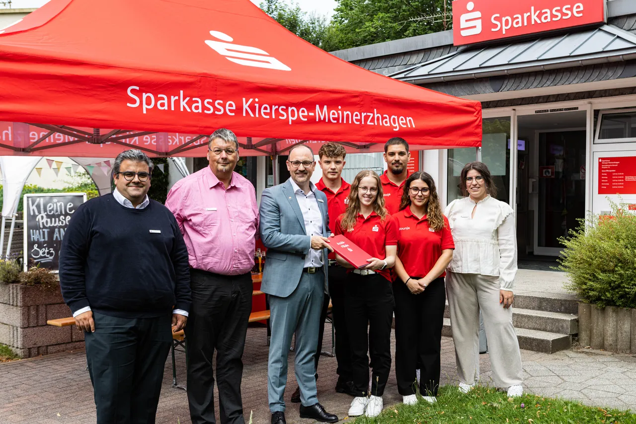 Manuel Jhuj, Markus Hölscher-Bartsch und Torsten Rath überreichen vier Auszubildenden Urkunden vor der Sparkasse Kierspe-Meinerzhagen. Die Gruppe steht unter einem roten Pavillon mit dem Sparkassen-Logo.