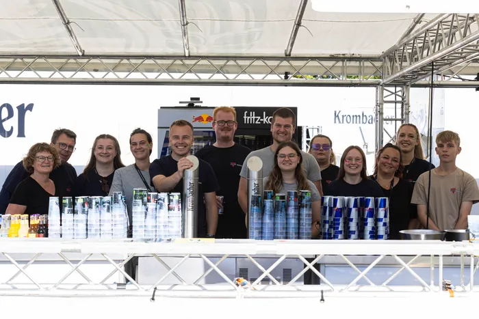 Eine Gruppe von Menschen steht lächelnd hinter einer Bar, die mit zahlreichen Getränkedosen und Flaschen bestückt ist.