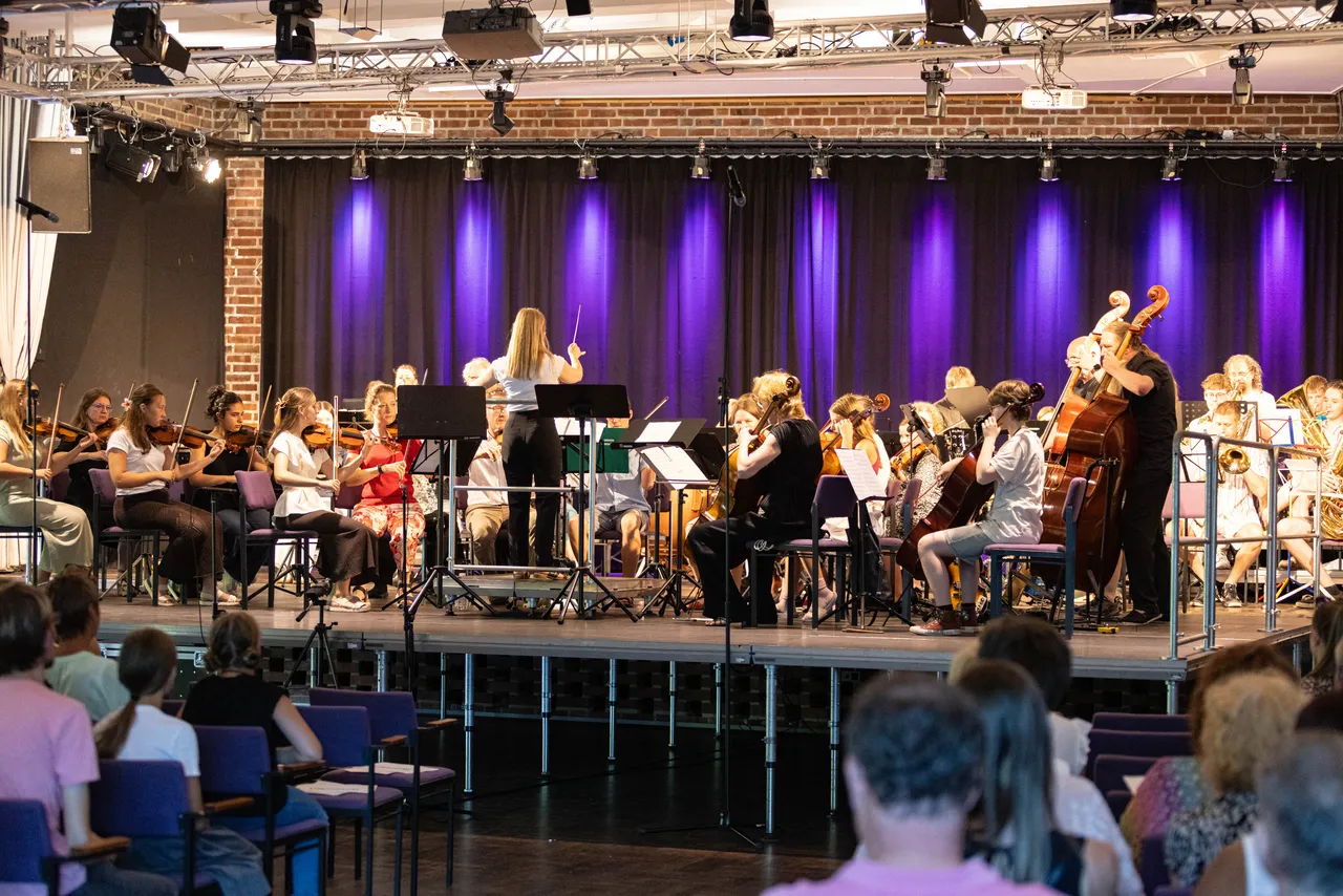 Ein großes Orchester spielt auf einer Bühne vor einem Publikum. Die Musikerinnen und Musiker sitzen auf Stühlen und spielen Geigen, Celli, Posaunen und andere Instrumente, während eine Dirigentin das Orchester leitet.