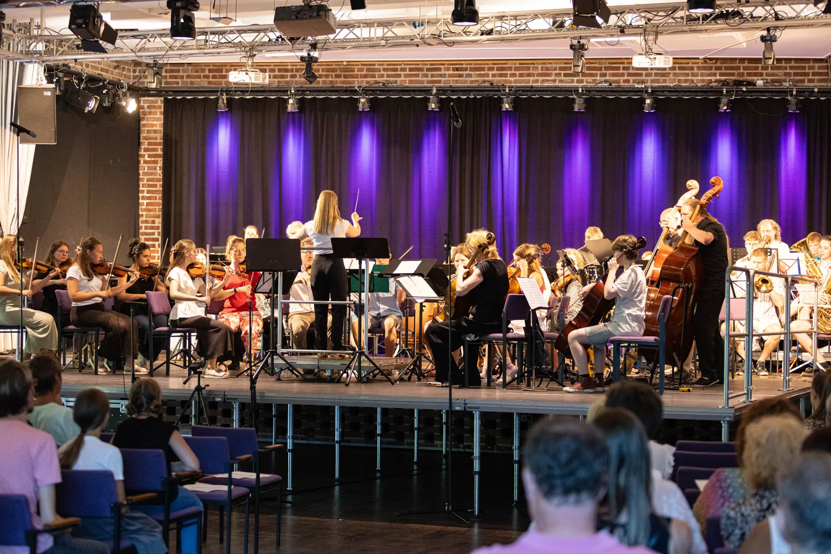 Ein großes Orchester spielt auf einer Bühne vor einem Publikum. Die Musikerinnen und Musiker sitzen auf Stühlen und spielen Geigen, Celli, Posaunen und andere Instrumente, während eine Dirigentin das Orchester leitet.