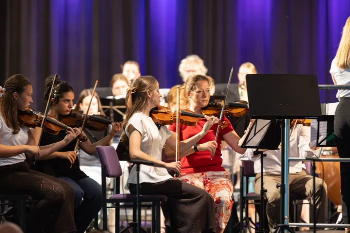 Ein Orchester spielt in einem Konzertsaal. Mehrere Musikerinnen und Musiker sitzen auf Stühlen und spielen Violine, während eine Dirigentin mit dem Rücken zum Betrachter zu sehen ist.