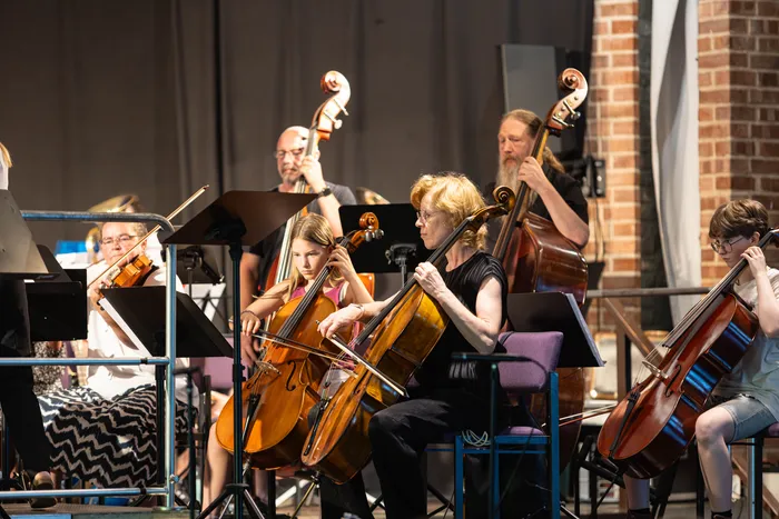 Ein Orchester spielt in einer Aula. Mehrere Musiker spielen Cello, während andere mit ihren Instrumenten im Hintergrund zu sehen sind.