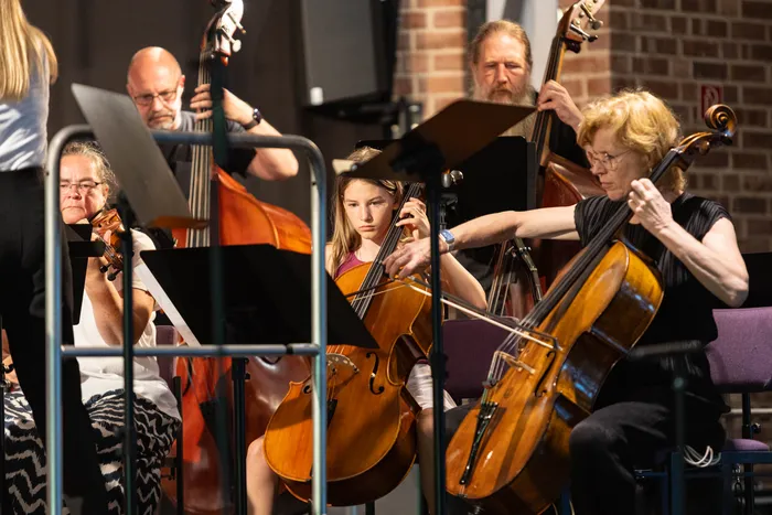 Ein Orchester spielt in einem Raum mit Backsteinwänden. Im Vordergrund eine junge Cellistin, umgeben von weiteren Musikern mit verschiedenen Instrumenten wie Geigen und Celli.