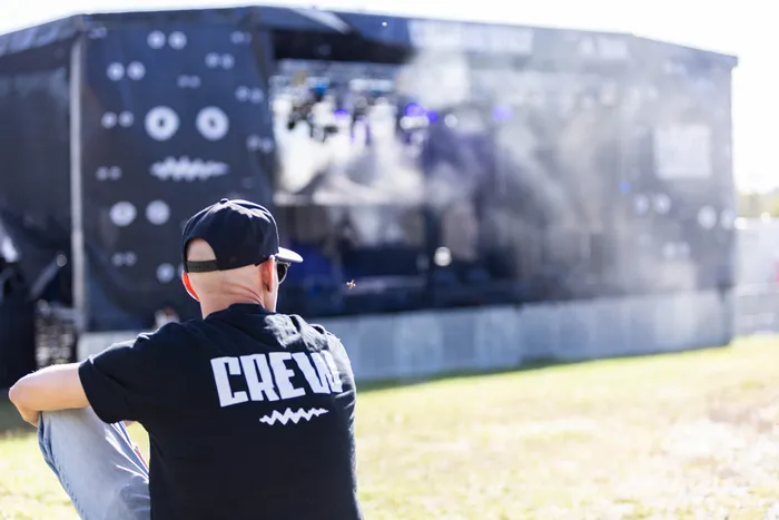 Ein Mann mit einem schwarzen „CREW“-T-Shirt sitzt auf einer Wiese und blickt auf eine Bühne beim Bautz-Festival.