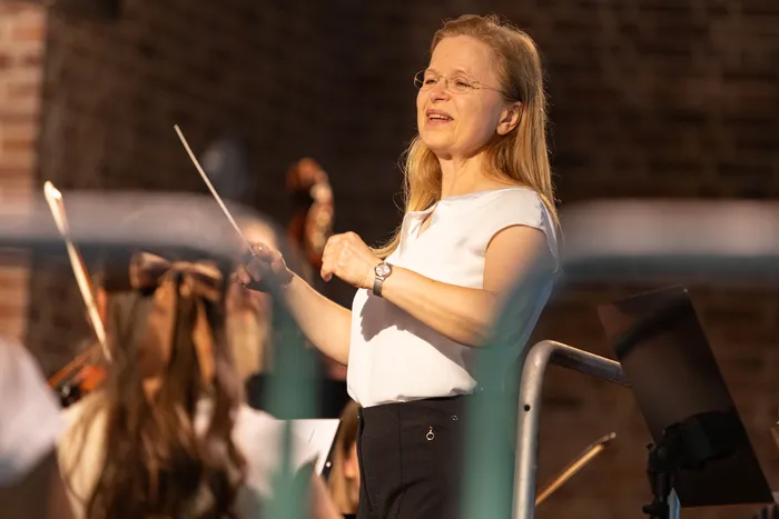 Cosima Breidenstein, Leiterin der Musikschule Volmetal, dirigiert das Sinfonieorchester mit einem Taktstock. Sie trägt ein weißes Oberteil und eine Brille. Im Hintergrund sind Musiker mit ihren Instrumenten zu sehen.