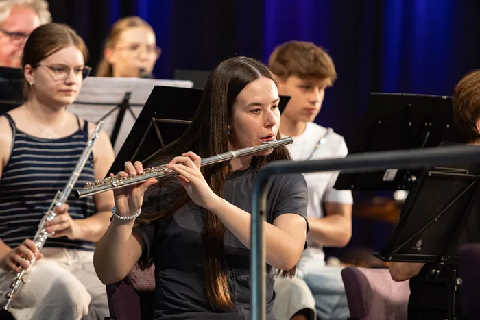 Eine junge Frau spielt Flöte in einem Orchester. Im Hintergrund sind weitere Musiker mit ihren Instrumenten und Notenständern zu sehen.