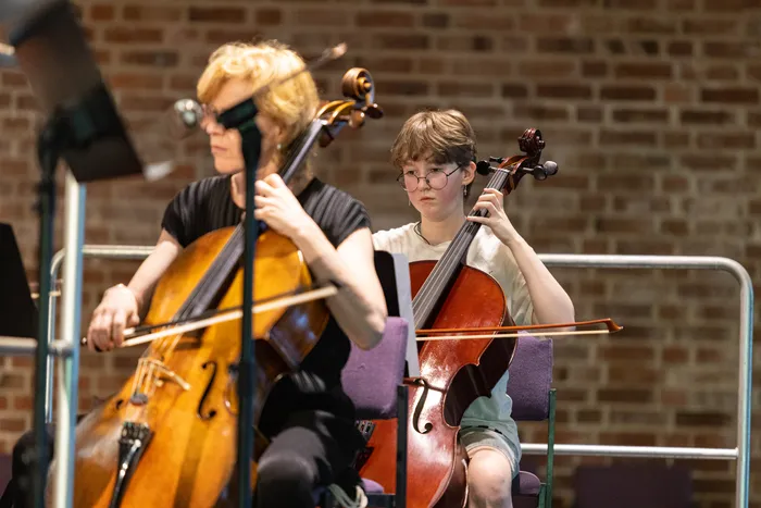 Zwei Cellistinnen spielen in einem Orchesterkonzert. Die jüngere Cellistin mit Brille konzentriert sich auf ihre Noten, während die ältere Cellistin den Bogen führt.