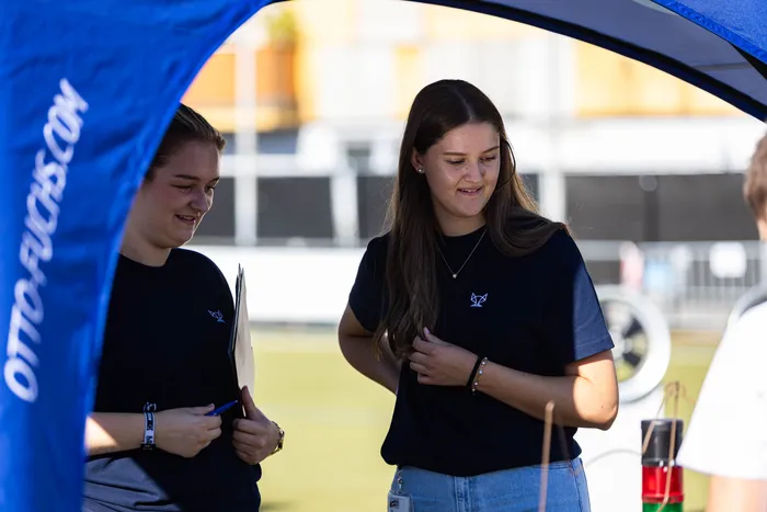 Zwei junge Frauen in dunkelblauen T-Shirts stehen unter einem blauen Zelt beim Bautz-Festival; eine von ihnen hält ein Klemmbrett.