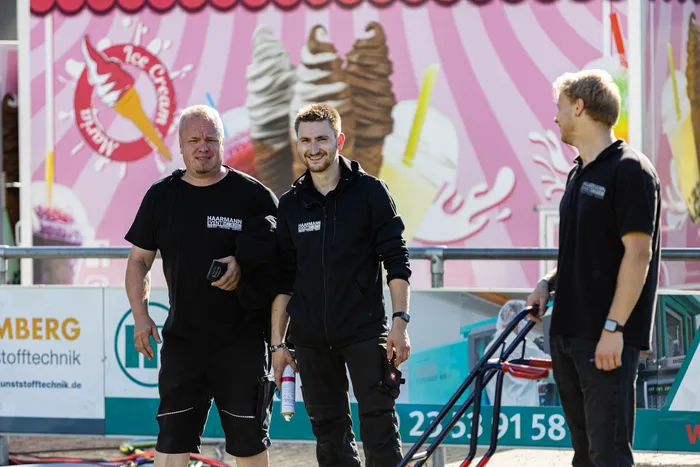 Drei Männer in schwarzer Arbeitskleidung stehen vor einer rosa-weiß gestreiften Kulisse mit Eiscreme-Motiven.