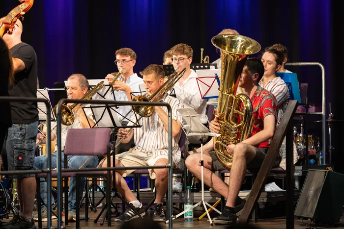 Ein Bläserensemble spielt auf einer Bühne. Mehrere Musiker sitzen auf Stühlen und spielen Posaunen, Trompeten und eine Tuba. Im Hintergrund ist ein Teil eines Schlagzeugs zu sehen.