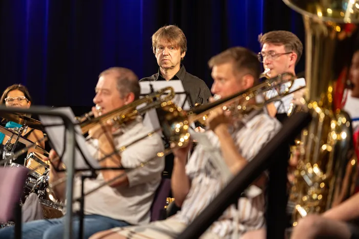 Ein Orchester spielt in einem Konzertsaal. Mehrere Musiker mit Blechblasinstrumenten sind im Vordergrund zu sehen, während ein Mann mit roten Haaren im Hintergrund steht.