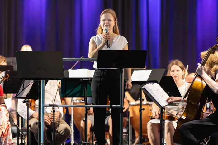 Cosima Breidenstein, Leiterin der Musikschule Volmetal, steht mit einem Mikrofon vor einem Orchester auf der Bühne. Musikerinnen und Musiker sitzen mit Notenständern und Instrumenten vor ihr.