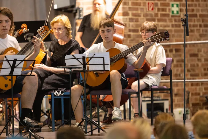 Ein Ensemble aus vier Gitarristen spielt in einem Konzertsaal. Die Musiker sitzen auf Stühlen vor Notenständern und konzentrieren sich auf ihr Spiel. Im Hintergrund sind weitere Musiker und eine Harfe zu sehen.