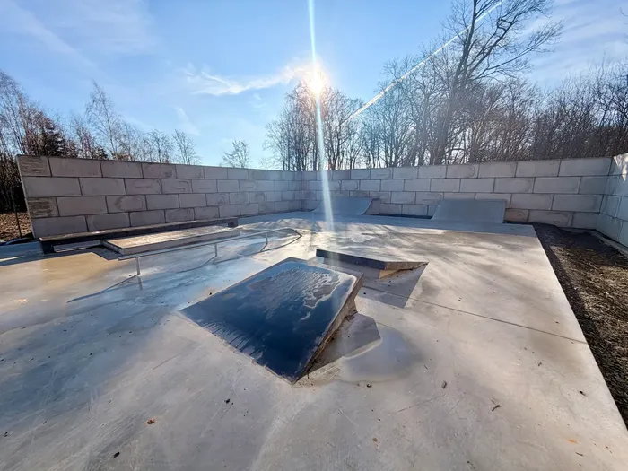 Betonflächen und Rampen einer modernen Skateranlage unter blauem Himmel mit kahlen Bäumen im Hintergrund.