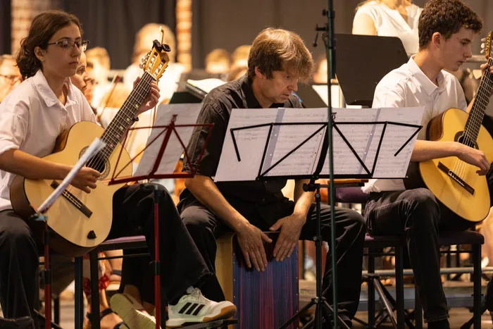 Drei Musiker spielen Gitarre und Percussion während eines Konzerts in der Aula eines Gymnasiums. Im Hintergrund sind weitere Musiker eines Orchesters zu sehen.