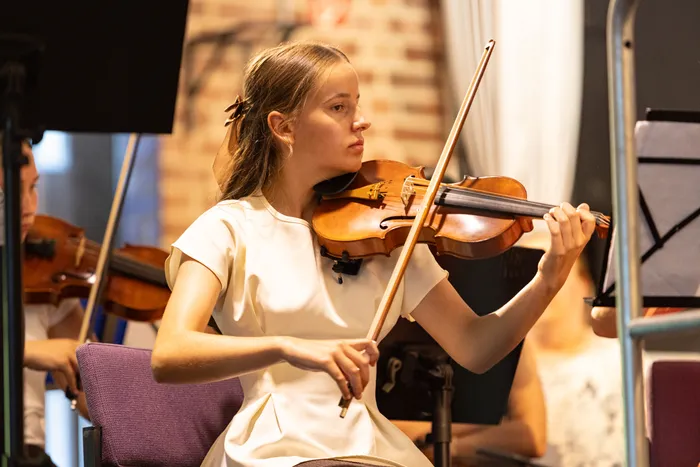 Eine junge Frau spielt konzentriert Violine während eines Konzerts. Im Hintergrund sind weitere Musiker und Notenständer zu sehen.