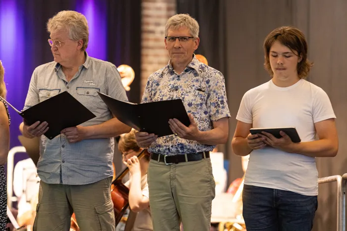 Drei Männer stehen nebeneinander und halten Notenblätter oder Tablets in den Händen. Im Hintergrund sind weitere Musiker und Instrumente eines Orchesters zu erkennen.