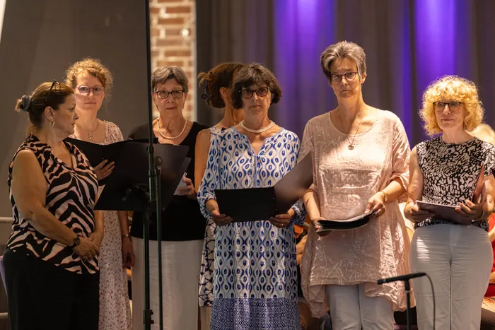 Sechs Frauen stehen auf einer Bühne und halten Notenblätter in den Händen. Sie scheinen zu singen oder zu proben, während sie in Richtung des Publikums blicken. Im Hintergrund ist eine Bühne mit violettem Licht zu sehen.