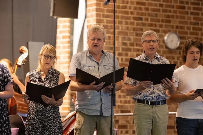 Chor singt in einem Innenraum. Vier Personen mit Notenblättern stehen im Vordergrund und singen, während im Hintergrund weitere Musiker und Instrumente zu sehen sind.