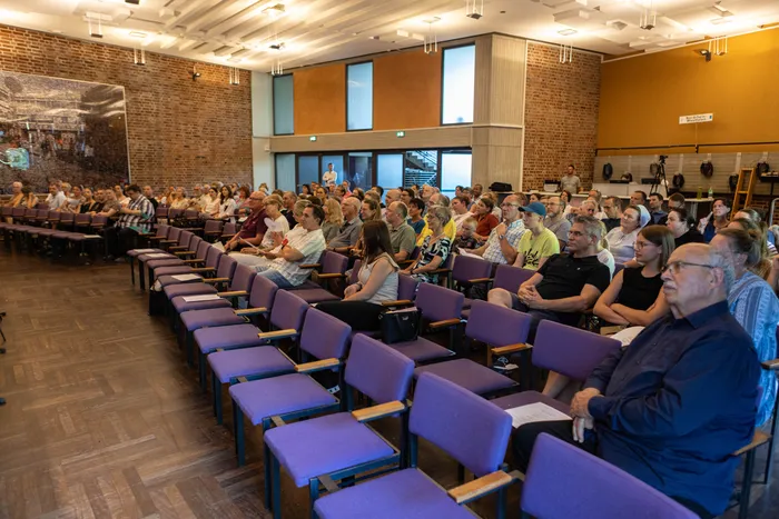 Ein Publikum sitzt auf violetten Stühlen in einer großen Aula und lauscht aufmerksam. Im Hintergrund befindet sich eine Bühne und ein Mann steht vor einer Tür.