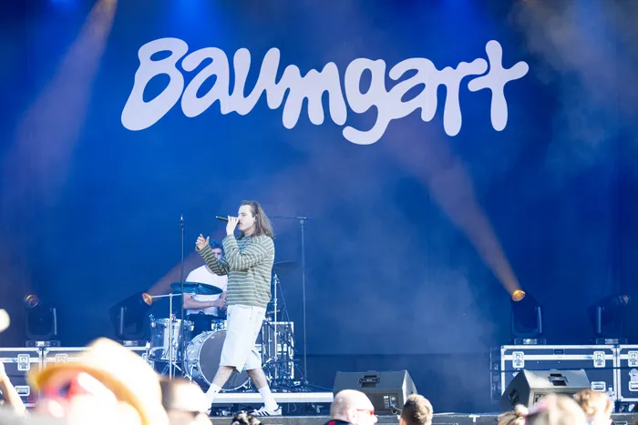 Ein Musiker singt in ein Mikrofon auf einer Bühne vor einem großen blauen Hintergrund mit dem weißen Schriftzug „Baumgart“.