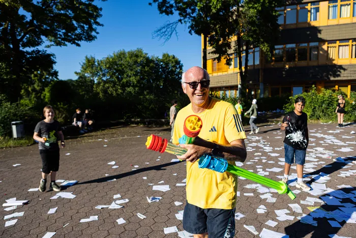 Ein Mann mittleren Alters mit Brille und gelbem T-Shirt hält eine Wasserpistole und lächelt in die Kamera. Im Hintergrund sind weitere Personen und ein Schulgebäude zu sehen. Papierstücke liegen auf dem Boden.