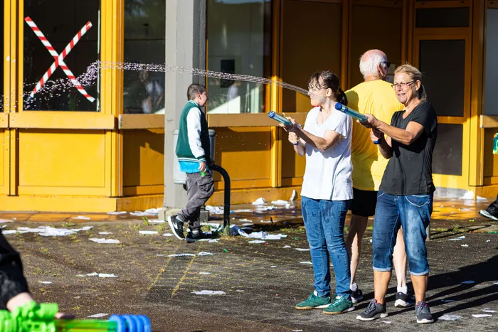 People spray each other with water guns in front of a yellow building with a red X on the windows.