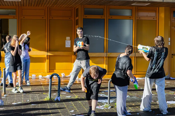 Die Schüler feiern mit Wasserpistolen und Konfetti vor dem Schulgebäude. Einige Schüler stehen, andere ducken sich, während sie mit Wasser bespritzt werden.