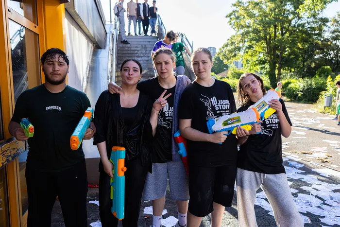 Fünf junge Menschen stehen mit Wasserpistolen in der Hand vor einer gelben Wand, einige sind mit Wasser bespritzt. Sie tragen schwarze T-Shirts mit Aufdruck und Shorts. Im Hintergrund sind weitere Personen und eine Treppe zu sehen.