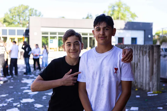Zwei lächelnde Jungen posieren vor der Kamera, der eine zeigt mit dem Finger auf sich selbst. Im Hintergrund sind weitere Schülerinnen und Schüler zu sehen.
