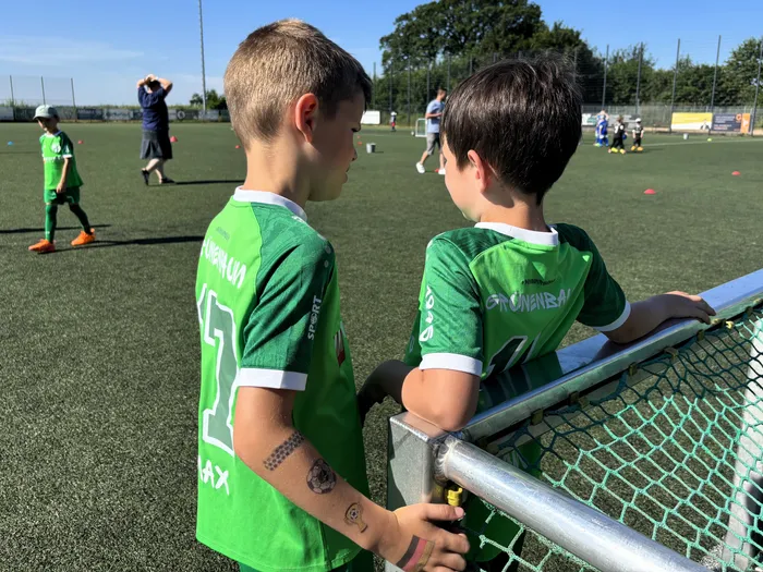 Zwei junge Fußballspieler mit den Trikotnummern 17 und 5 stehen mit dem Rücken zum Betrachter an der Seitenlinie eines Fußballfeldes. Im Hintergrund trainieren weitere Spieler.