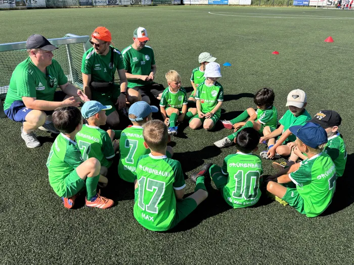 Fußballtrainer und Kinder sitzen im Kreis auf dem grünen Kunstrasen und besprechen Taktiken. Die Kinder tragen grüne Trikots mit der Aufschrift 