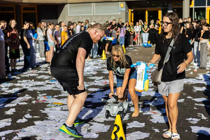 Drei Personen sind inmitten einer Wasserschlacht auf dem Schulhof der Rothensteinschule zu sehen. Ein Mann und eine Frau bespritzen ein Kind auf einem Tretroller mit Wasserpistolen, während im Hintergrund eine Menschenmenge zusieht.