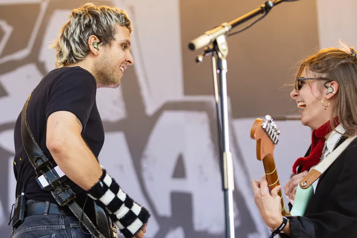 Paula Carolina mit einer hellblauen E-Gitarre und ein Mann mit schwarz-weißem Armwärmer lachen sich auf der Bühne des Bautz-Festivals an.