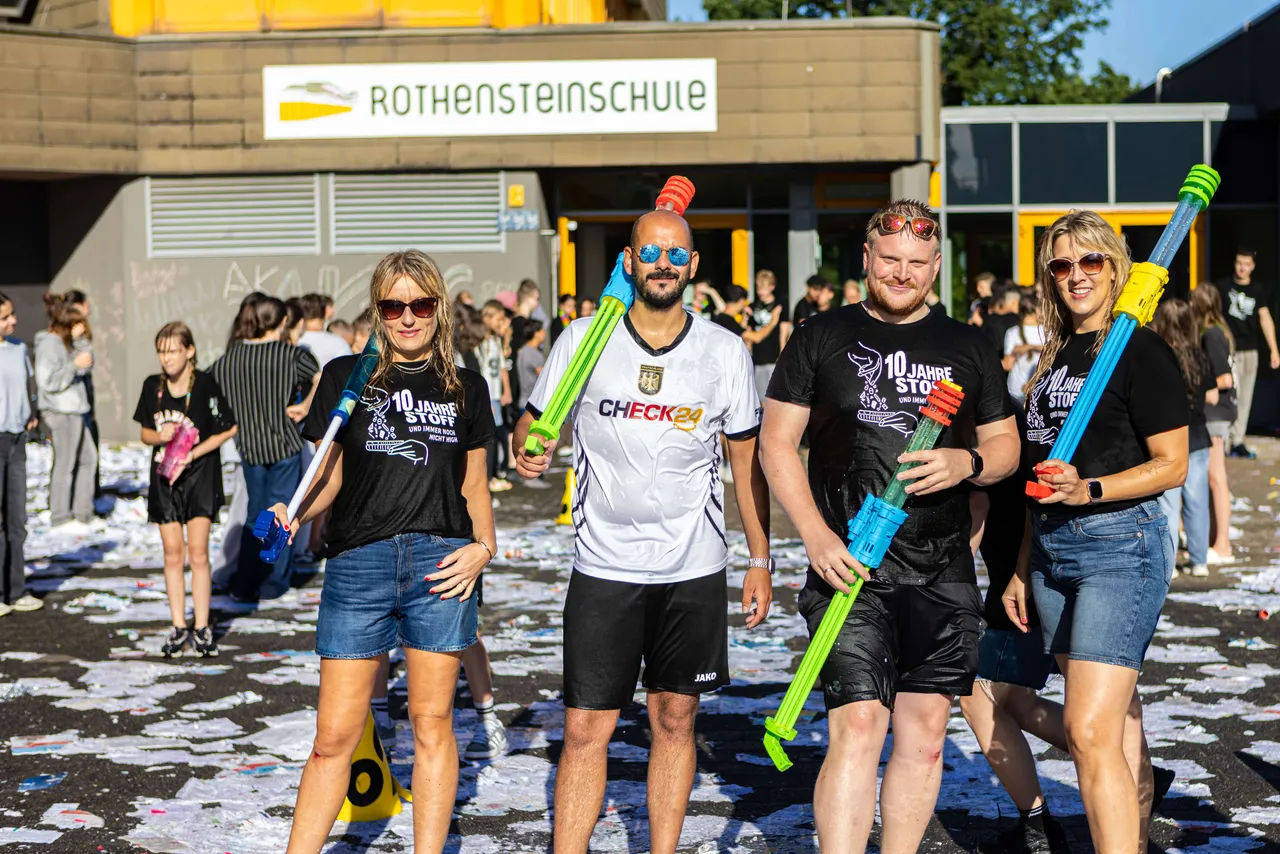 Eine Gruppe von Menschen schreitet vor einer Schule mit Wasserpistolen bewaffnet durch Konfetti. Sie tragen T-Shirts mit Aufdruck und Jeansshorts.
