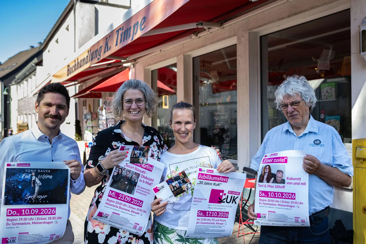 Vier Personen stehen lächelnd vor einer Buchhandlung und halten Programmhefte für die Veranstaltungsreihe KuK in der Hand. Im Hintergrund ist ein belebter Platz zu sehen.