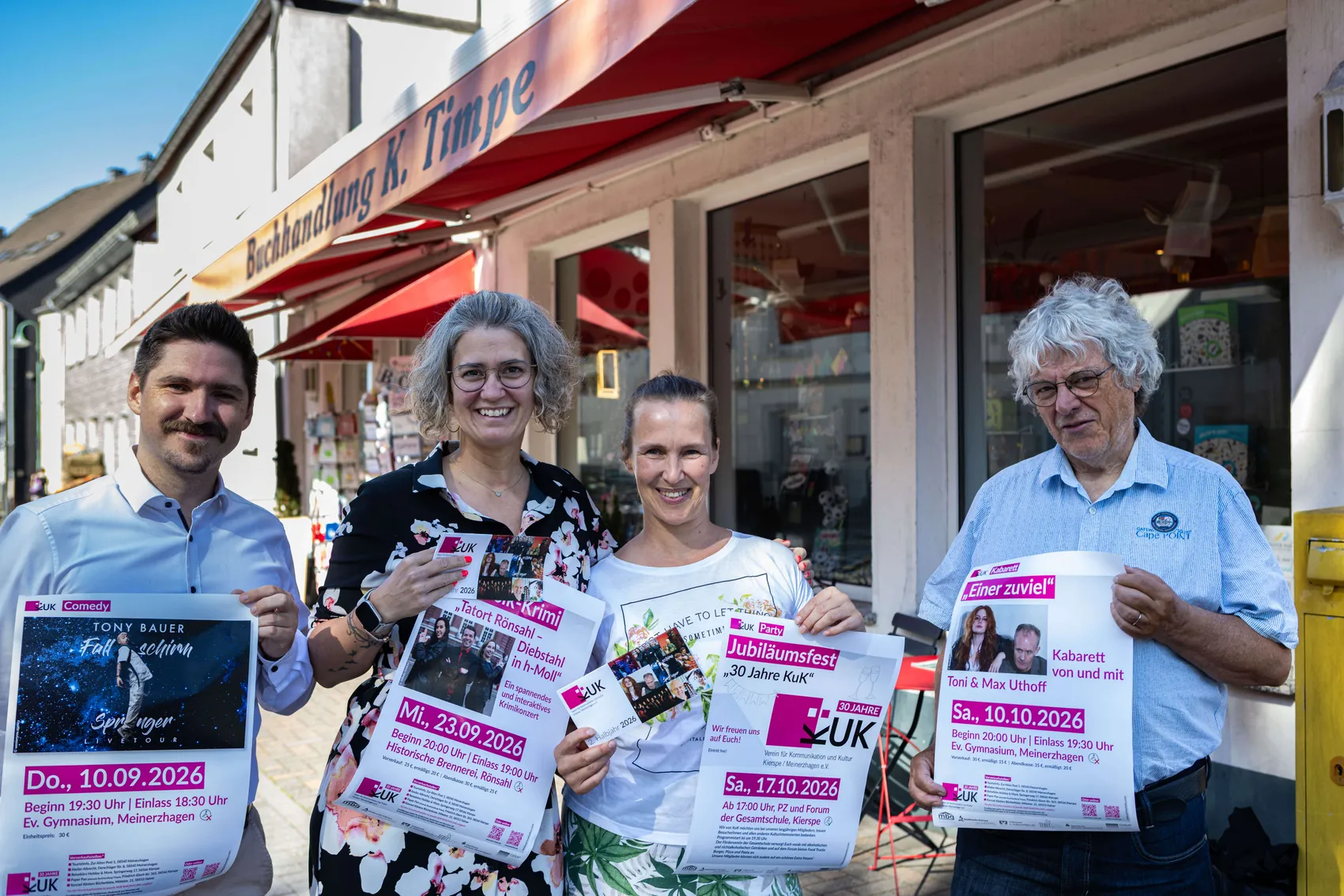 Vier Personen stehen lächelnd vor einer Buchhandlung und halten Programmhefte für die Veranstaltungsreihe KuK in der Hand. Im Hintergrund ist ein belebter Platz zu sehen.