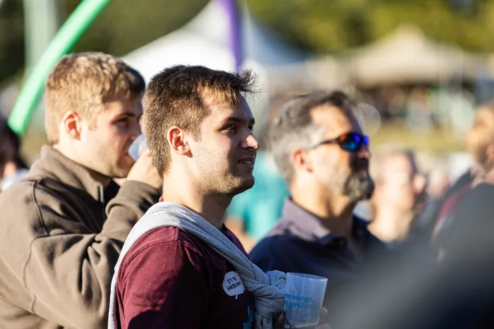 Ein junger Mann mit weinrotem T-Shirt und grauem Pullover um die Schultern blickt lächelnd zur Seite und hält einen Becher.