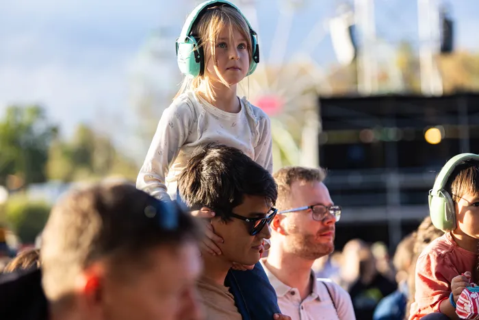 Ein kleines Mädchen mit mintgrünen Kopfhörern sitzt auf den Schultern eines Mannes bei einem Musikfestival.