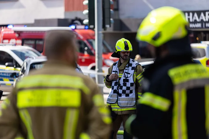 Ein Feuerwehrmann mit gelber Schutzkleidung und Helm steht vor Einsatzfahrzeugen und erklärt etwas. Im Hintergrund sind weitere Einsatzkräfte und ein Gebäude zu sehen.
