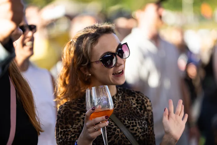 Eine lächelnde Frau mit Sonnenbrille und Leopardenmuster-Oberteil hält ein Glas Aperol Spritz in einer Menschenmenge.