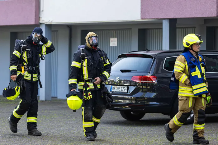 Drei Feuerwehrleute in voller Montur stehen vor einem schwarzen PKW mit dem Kennzeichen MK NB 1724. Im Hintergrund ist ein Gebäude mit grauen Türen zu sehen.