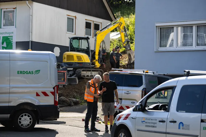 Zwei Arbeiter mit Warnwesten stehen vor einem Bagger und mehreren Transportern mit dem Aufdruck „ERDGAS“ an einer Baustelle vor einem Wohnhaus.