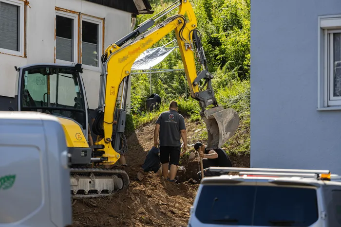 Ein Bagger arbeitet auf einer Baustelle vor Häusern, während ein Bauarbeiter im Hintergrund einen Hang hinaufgeht.